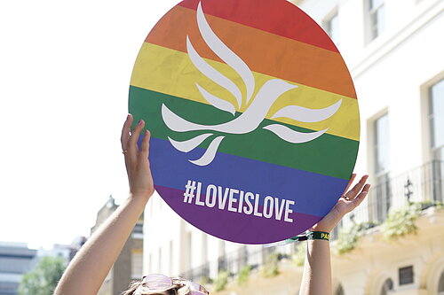 Young person holding a round sign over their head with a rainbow-striped, pride flag design overlaid with the Liberal Democrat bird of liberty emblem and #LoveIsLove