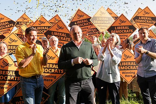 Ed Davey and the Liberal Democrats celebrate local election wins. Ed Davey, Charlie Maynard, Olly Glover and Liz Leffman stand in front of a crowd with Lib Dem diamond signs as golden confetti rains down.